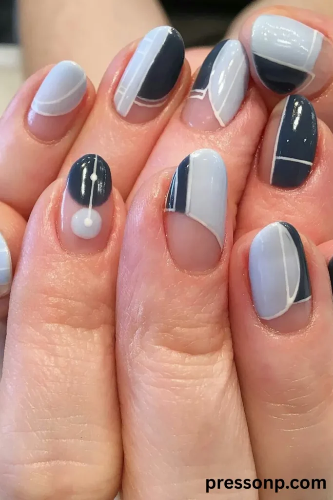A close-up of hands displaying a geometric negative-space manicure on short oval nails, featuring abstract color-blocked sections of slate gray and pale ice blue outlined with crisp white lines over a bare, natural nail base.