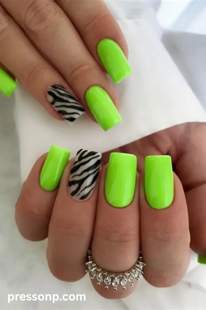Close-up of long square-shaped nails painted in a glossy, high-visibility neon lime green polish, accented by a striking black and white zebra stripe pattern on the ring finger.