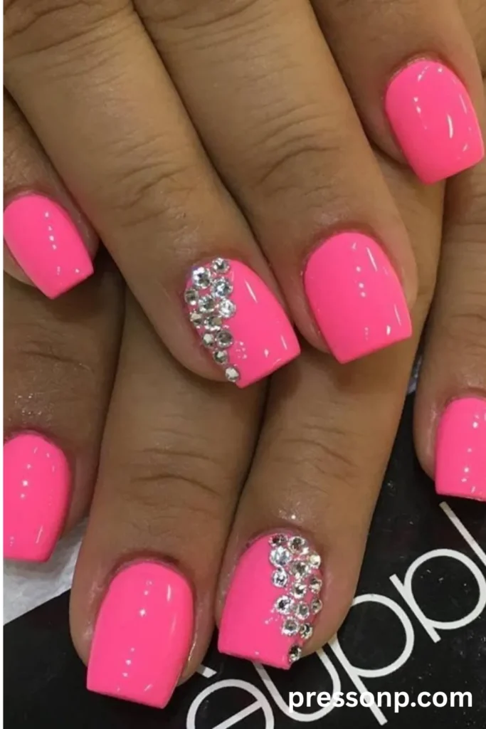 Close-up of short square nails coated in a high-gloss neon hot pink polish, featuring sparkly silver rhinestone clusters on the ring finger accent nails.
