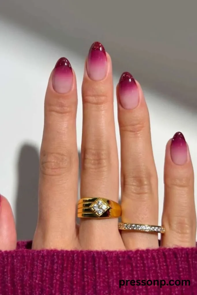A close-up of a hand showcasing an elegant ombré manicure on medium-length oval nails, featuring a seamless fade from a sheer nude base into a deep magenta-pink, capped with shimmering maroon-glitter tips.
