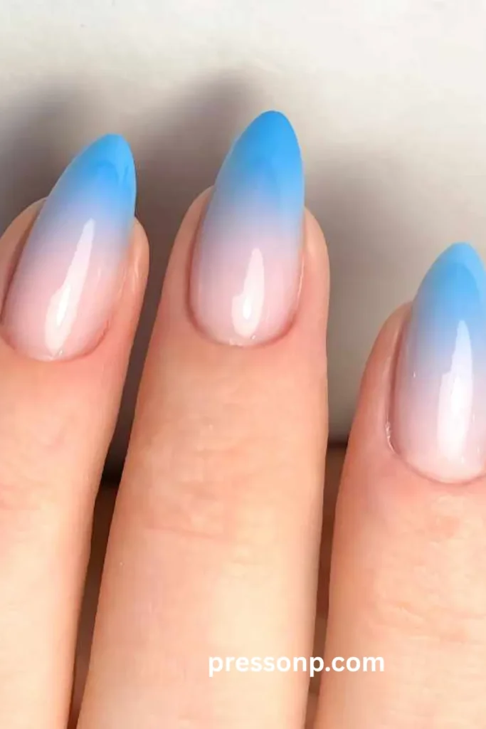 A close-up photograph of a hand showcasing a seamless gradient ombré manicure on long almond nails, transitioning smoothly from a soft, sheer milky pink at the cuticles to a vibrant, sky-blue color at the tips.