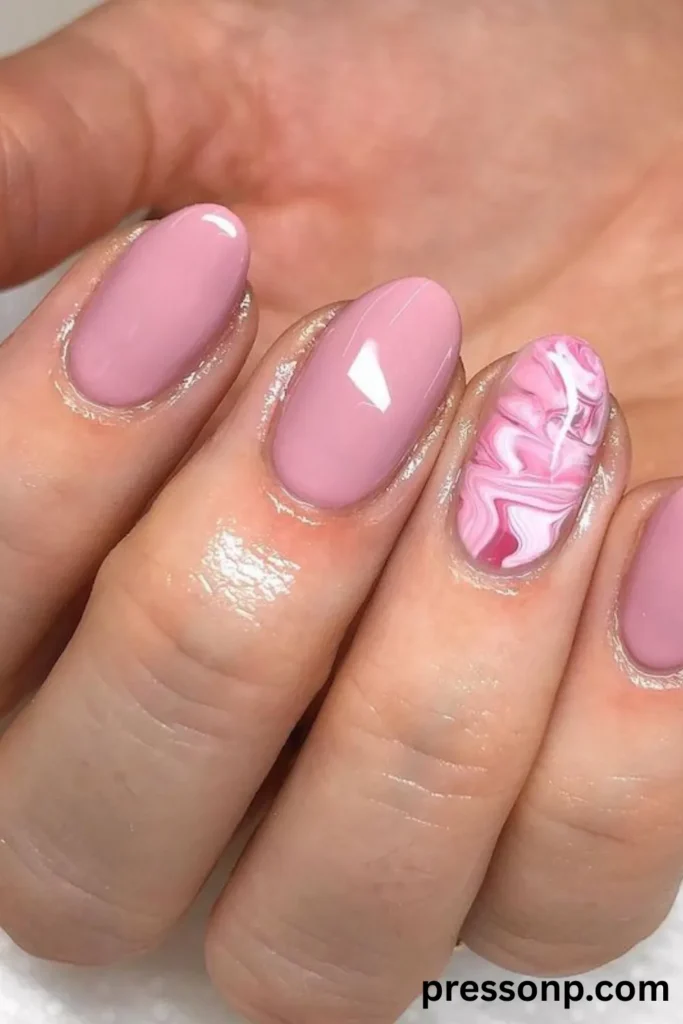 A close-up of a hand displaying a glossy mauve-pink manicure on short, natural oval nails, featuring a single accent ring finger decorated with a fluid, multi-toned pink and white marble swirl design.