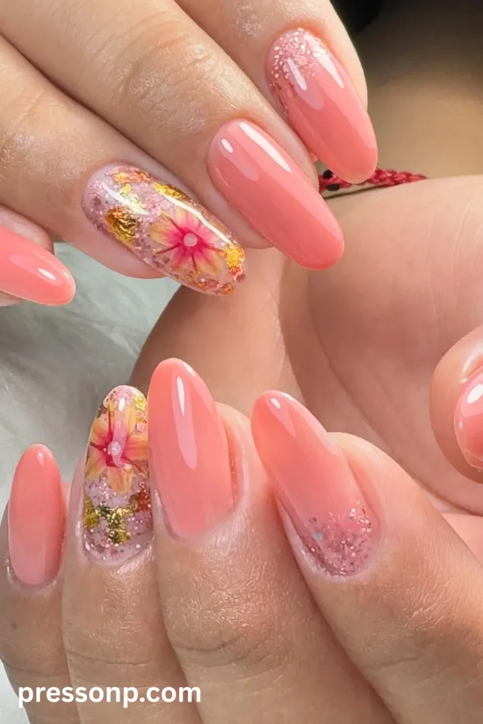 Close-up of long almond-shaped nails coated in a glossy pastel peach polish, featuring clear accent nails encapsulated with dried pink flowers, gold foil, and pink glitter.
