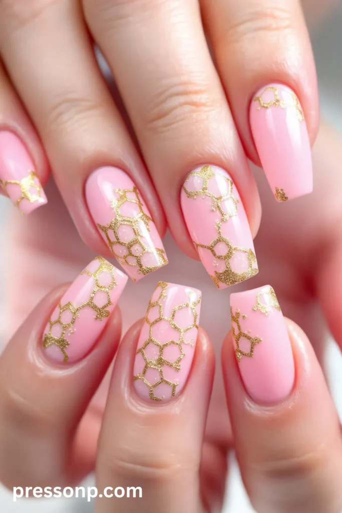 Close-up of long coffin-shaped nails featuring a glossy baby pink base adorned with an intricate, shimmering gold honeycomb pattern.