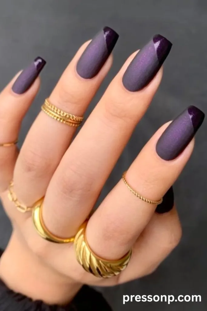 Close-up of long coffin-shaped nails featuring a deep plum matte base with a velvet metallic sheen, finished with sharp, high-gloss French tips and styled with statement gold rings