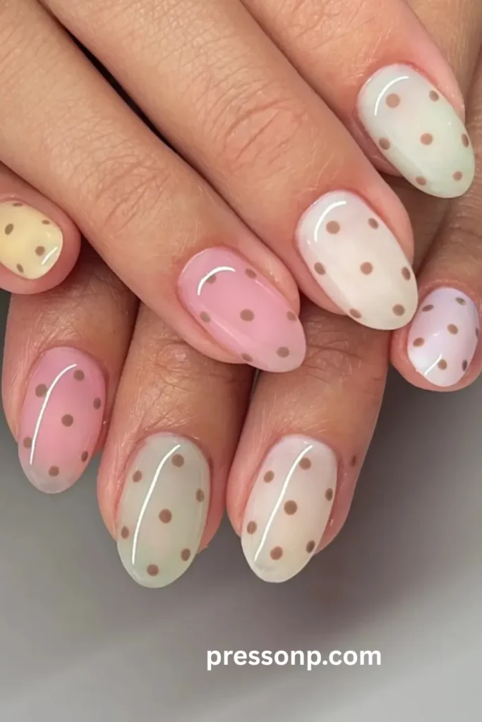 A close-up photograph of two hands interlaced to showcase a soft, pastel skittle manicure on short oval nails, uniformly covered in a minimalist pattern of small, evenly spaced brown polka dots over sheer jelly base colors.