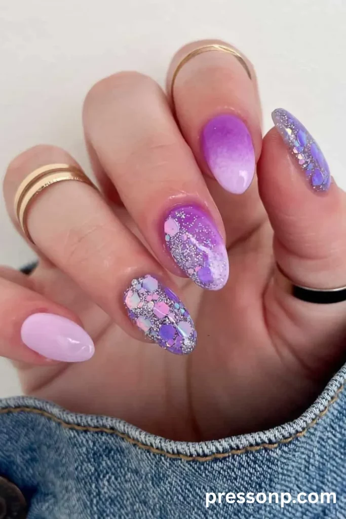 Close-up of medium almond-shaped nails featuring a mix of a purple-to-lavender ombré gradient, a solid pastel lilac nail, and heavy iridescent silver and purple glitter tips.