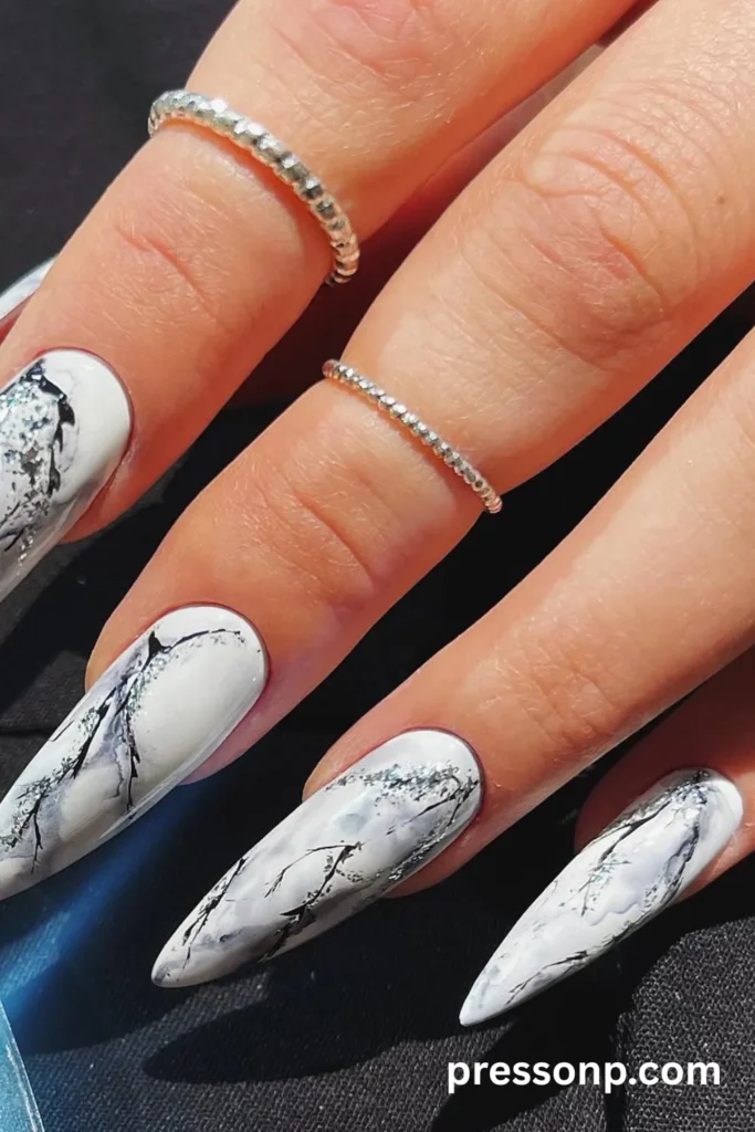 A close-up photograph of a hand displaying a luxurious Carrara marble-inspired manicure on long stiletto nails, featuring a crisp white base intricately detailed with realistic gray veins, smoky black swirls, and subtle hints of shimmering silver glitter.