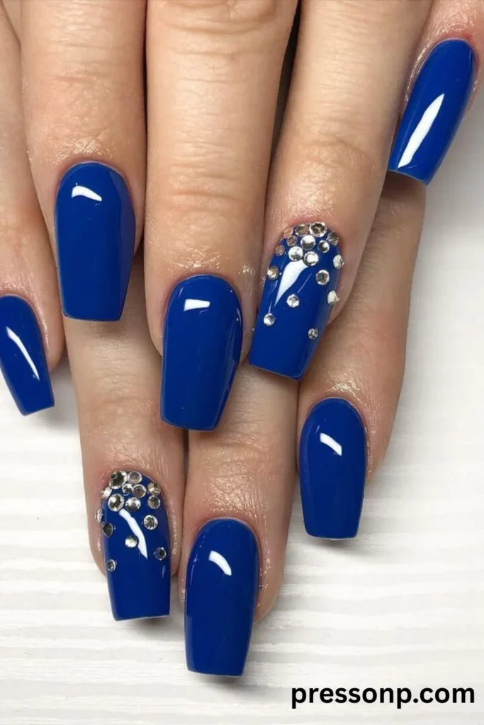 Close-up of long coffin-shaped nails painted in a highly glossy, solid royal blue polish, featuring accent nails decorated with sparkling silver rhinestones clustered near the cuticles.
