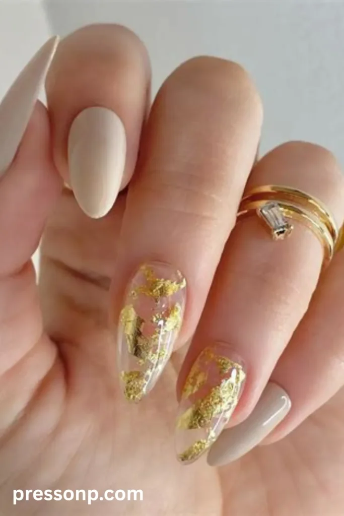 Close-up of almond-shaped nails featuring a mix of solid matte nude-beige polish and clear accent nails decorated with abstract metallic gold foil flakes.
