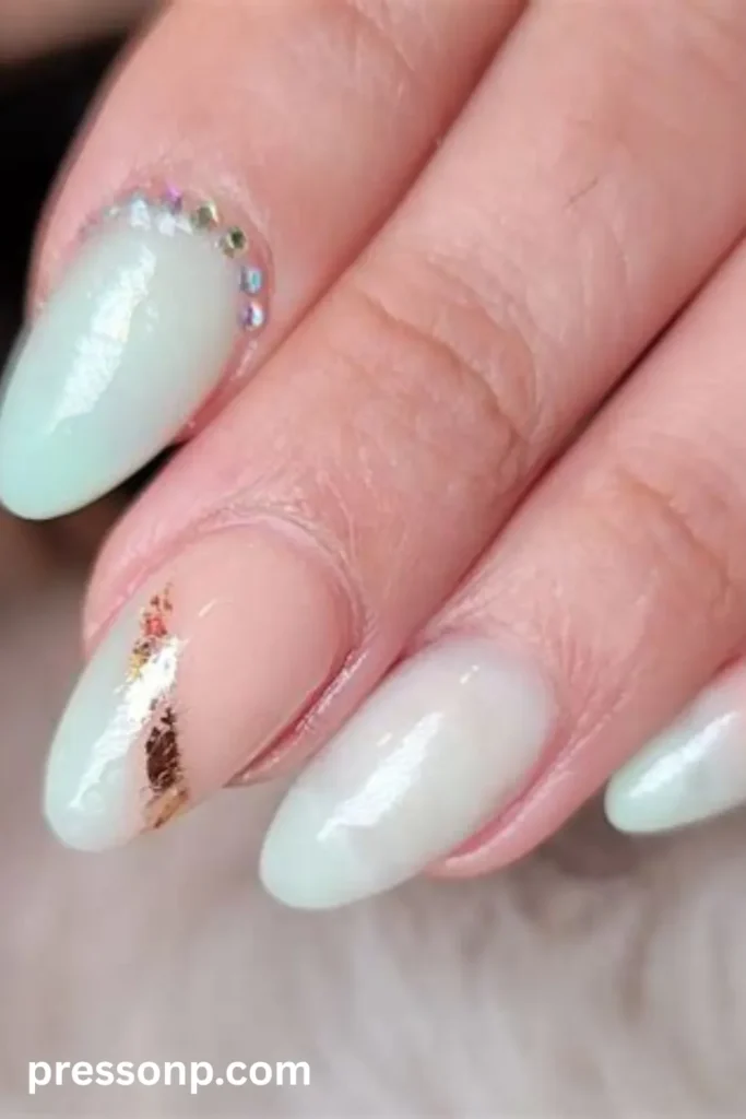 Close-up of long oval nails featuring a pale seafoam green polish, a curved row of iridescent rhinestones along the cuticle line, and a nude accent nail bordered with gold foil.