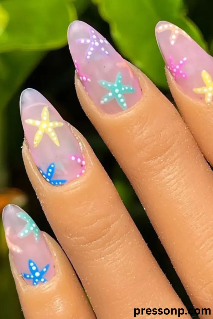 A close-up of long, almond-pointed extensions showcasing a whimsical summer manicure with a translucent, matte sea-glass base, decorated with colorful hand-painted pastel starfish detailed with white tiny dots.