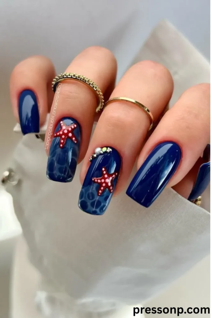 A close-up of a hand showcasing medium-length, square-shaped nails with a high-gloss deep navy blue manicure, featuring clear water-marbled accent nails decorated with hand-painted red starfish and tiny rhinestone cuticles.