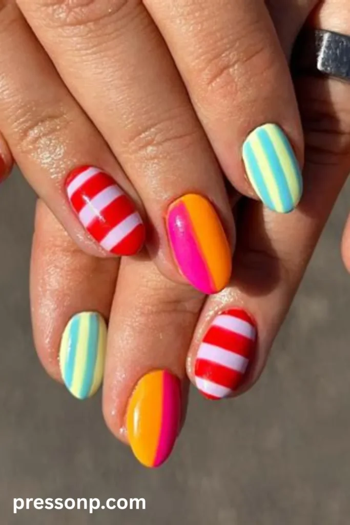 Close-up of hands displaying short oval nails painted in a bright mix of red and lilac horizontal stripes, yellow and blue vertical stripes, and pink and orange split color blocking.