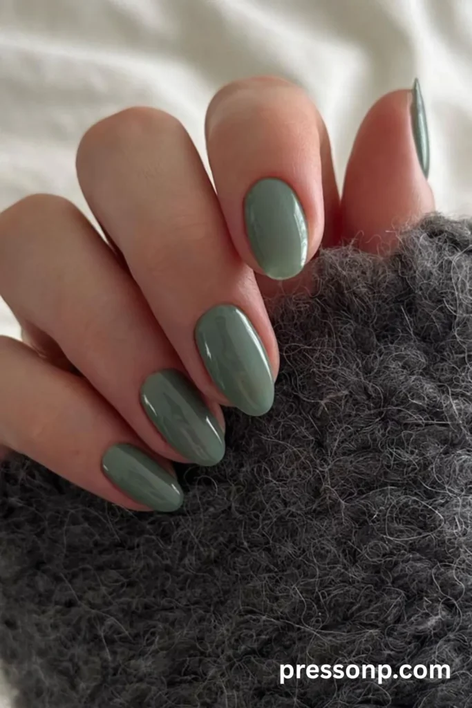 A close-up of a hand displaying a solid, minimalist sage olive green manicure on medium oval nails, beautifully contrasted against a textured, dark gray knit sweater.