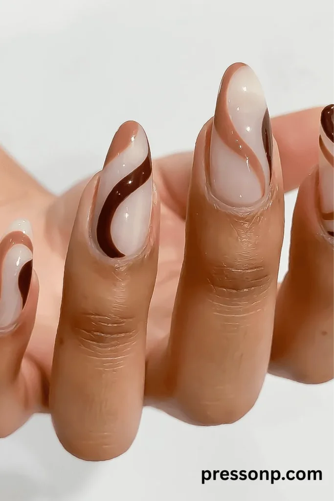 A close-up of long, almond-shaped manicured nails featuring an abstract swirl design in shades of espresso brown, iced coffee beige, and creamy white over a neutral base.