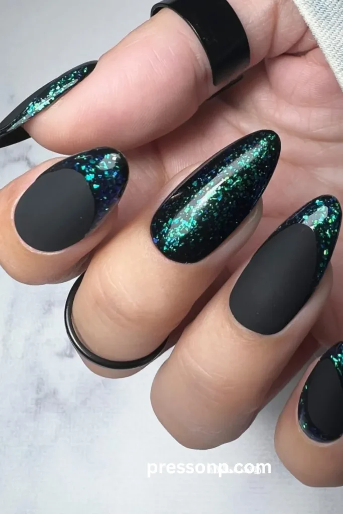 A close-up photograph of a hand showcasing a dramatic black and green manicure on long almond nails, seamlessly blending ultra-flat matte black finishes with a high-gloss, shimmering emerald and teal glitter flake base.