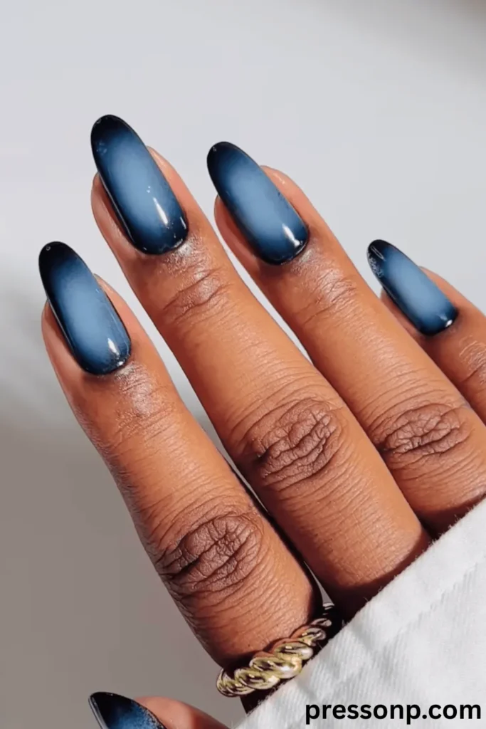A close-up of a hand with deep skin tones displaying long, oval-shaped nails with a high-gloss velvet aura manicure that transitions from midnight black edges to a glowing, metallic slate-blue center.