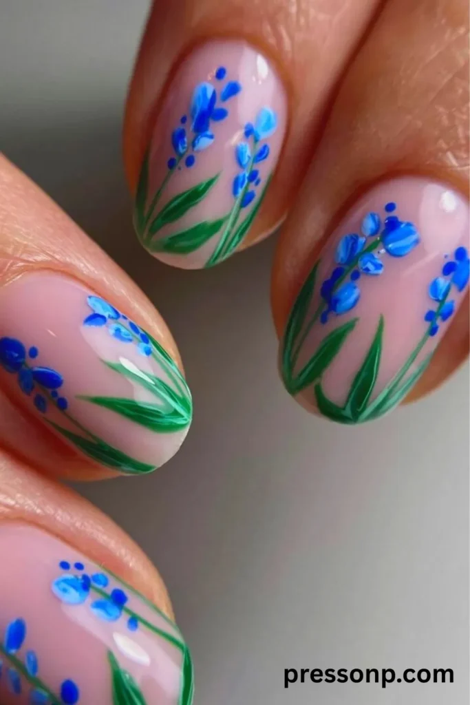 A close-up of a hand showcasing a high-gloss spring manicure on short oval nails, featuring a sheer pale-pink base delicately hand-painted with vibrant blue wildflower stems and green leaves.