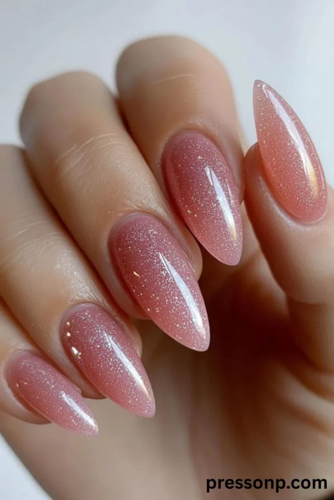A close-up of a hand showcasing a clean, elegant manicure on long almond-shaped nails, featuring a semi-translucent rose-pink gel base infused with fine silver and gold micro-shimmer glitter.
