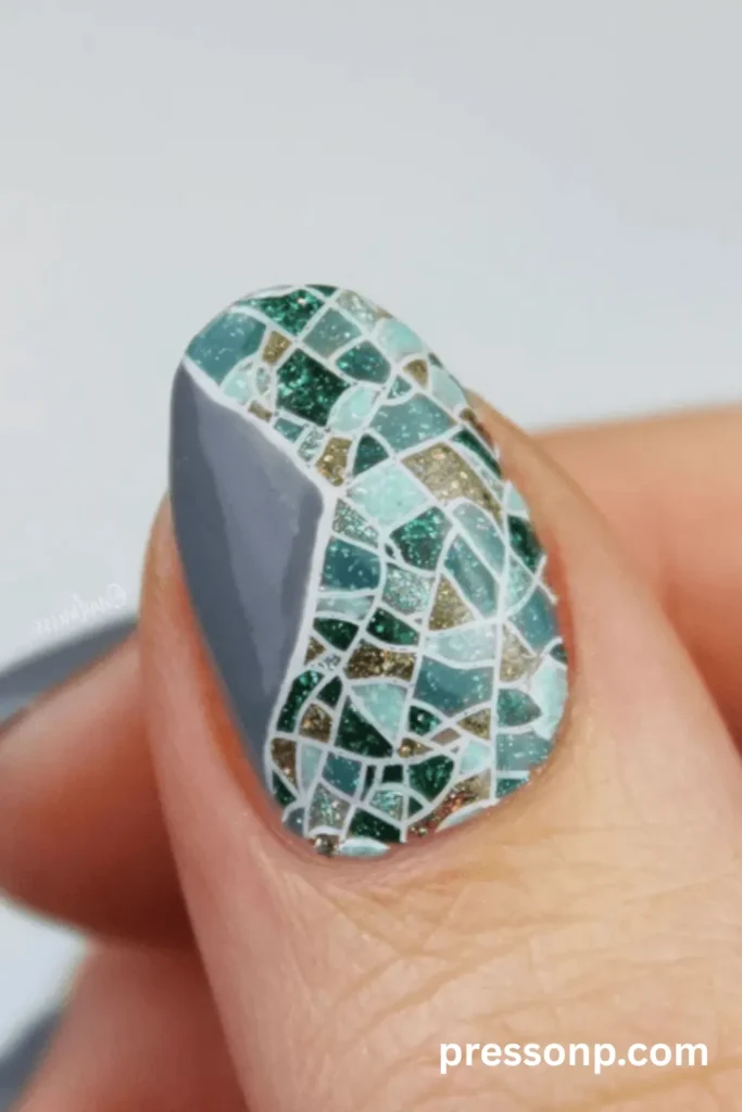 An extreme macro close-up of a short oval nail featuring an intricate stained glass mosaic manicure. The design combines a solid gray negative space block with an array of shimmering green, teal, and gold glitter "tiles" separated by crisp white grout lines.