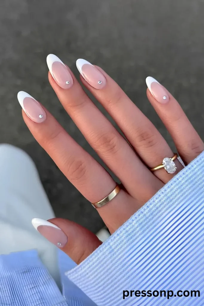 Close-up of long almond-shaped nails featuring a classic white French tip manicure, subtly accented with a single sparkling crystal rhinestone centered near each cuticle.
