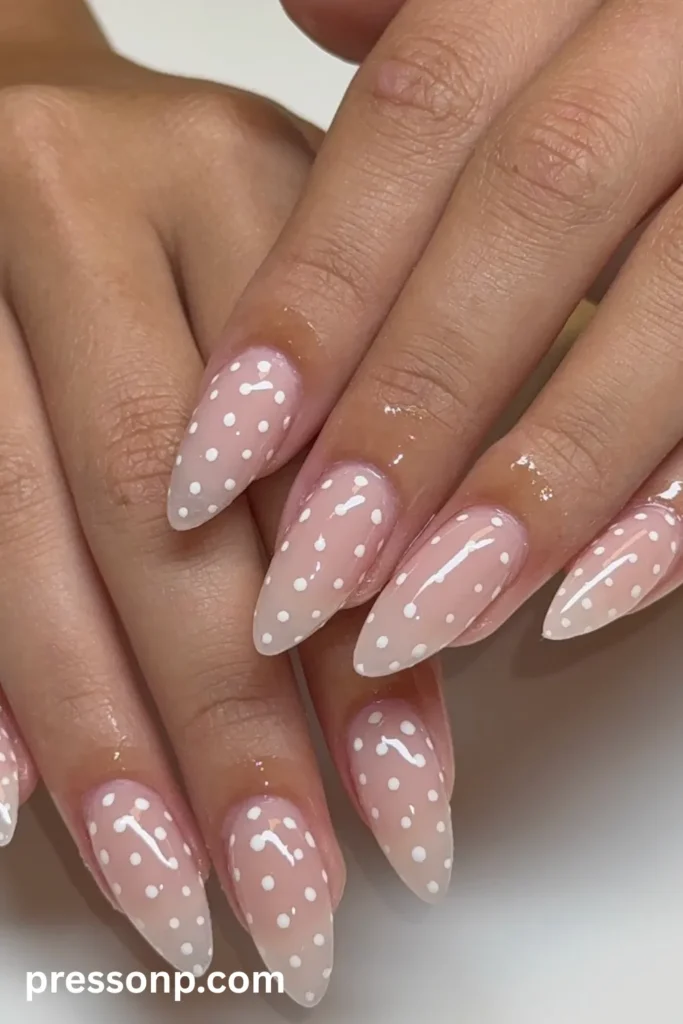 Close-up of long almond-shaped nails featuring a translucent, soft coral-pink base decorated with a scattered pattern of small white polka dots and a glossy finish.