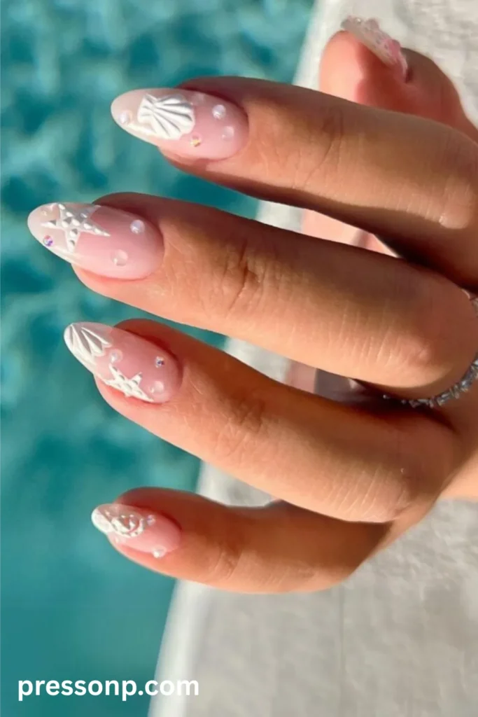 Close-up of almond-shaped nails on a sheer pink base, decorated with 3D white textured seashell and starfish charms alongside tiny scattered pearls.