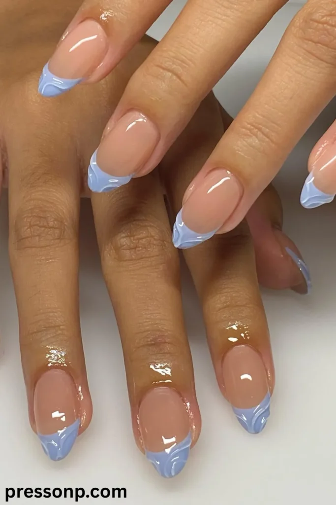A top-down close-up of a manicure featuring medium almond-shaped nails with a high-gloss nude base and periwinkle blue French tips detailed with a subtle 3D marbled or textured swirl pattern.