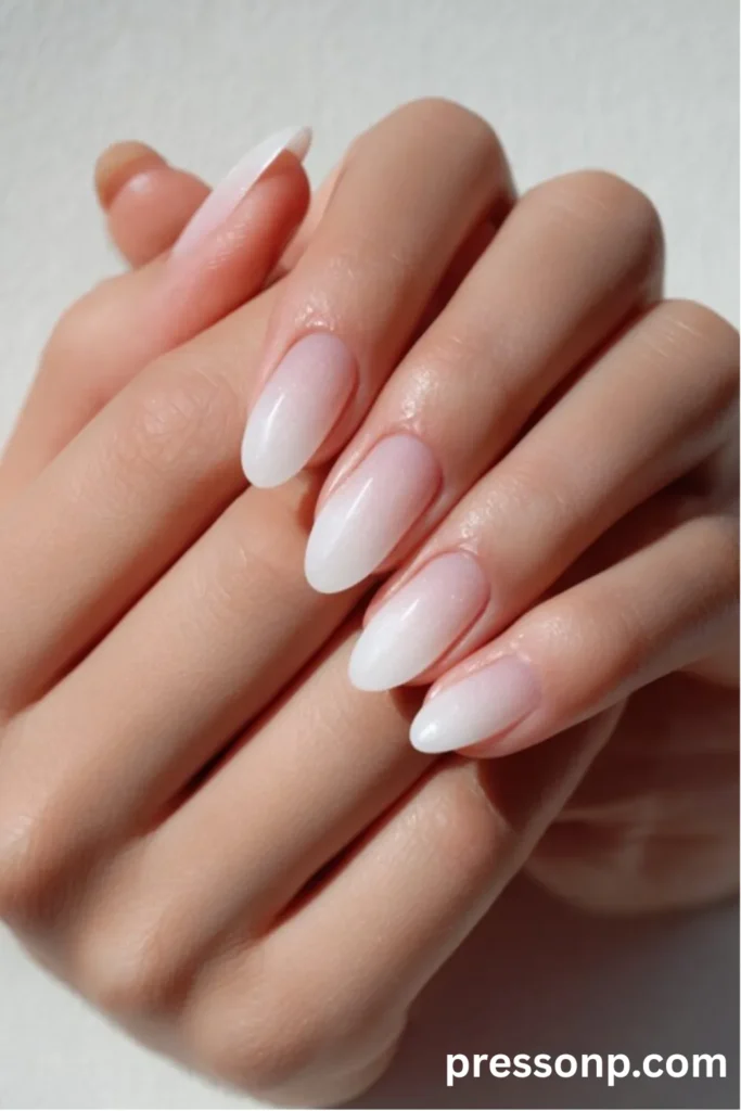 Close-up of hands with elegant almond-shaped nails featuring a soft milky white boomer ombré gradient that fades seamlessly from a sheer nude base to soft white tips.