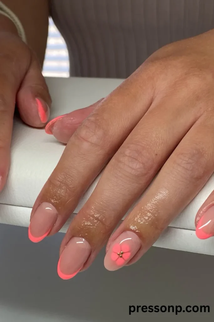 Close-up of hands displaying an oval-shaped manicure with bright coral French tips on a sheer nude base, featuring a simple painted coral flower accent nail.