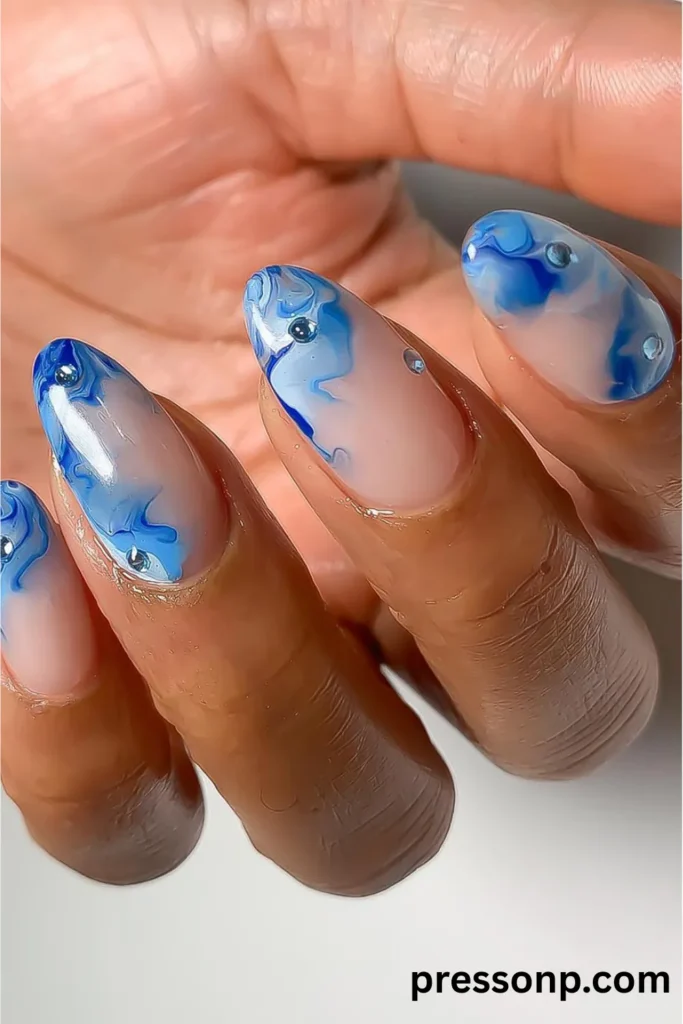 Close-up of long almond-shaped nails featuring a sheer nude base with fluid blue marble swirls and clear 3D water droplet accents.
