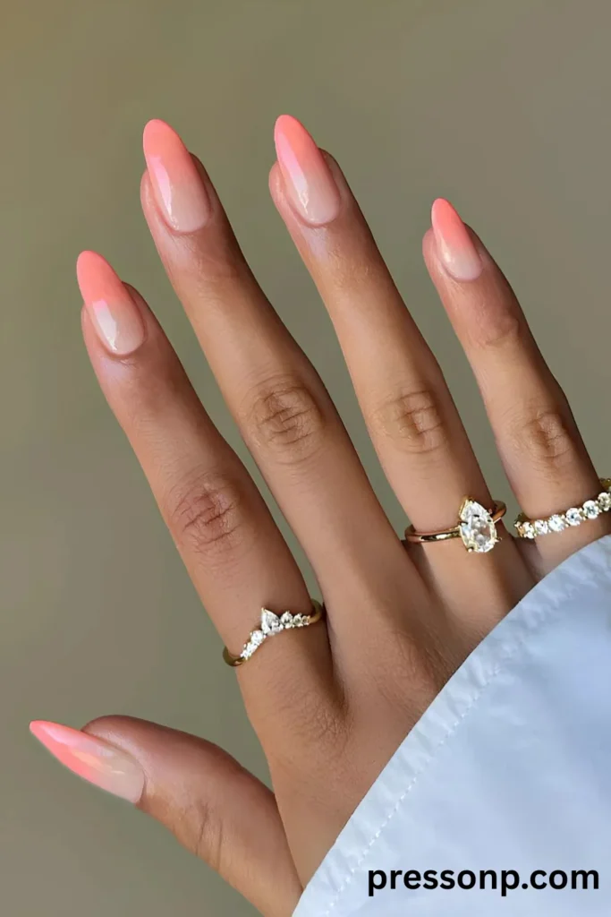 Close-up of a hand with long almond-shaped nails featuring a soft ombré gradient that transitions from a sheer nude base to vibrant coral pink tips, accessorized with gold rings.