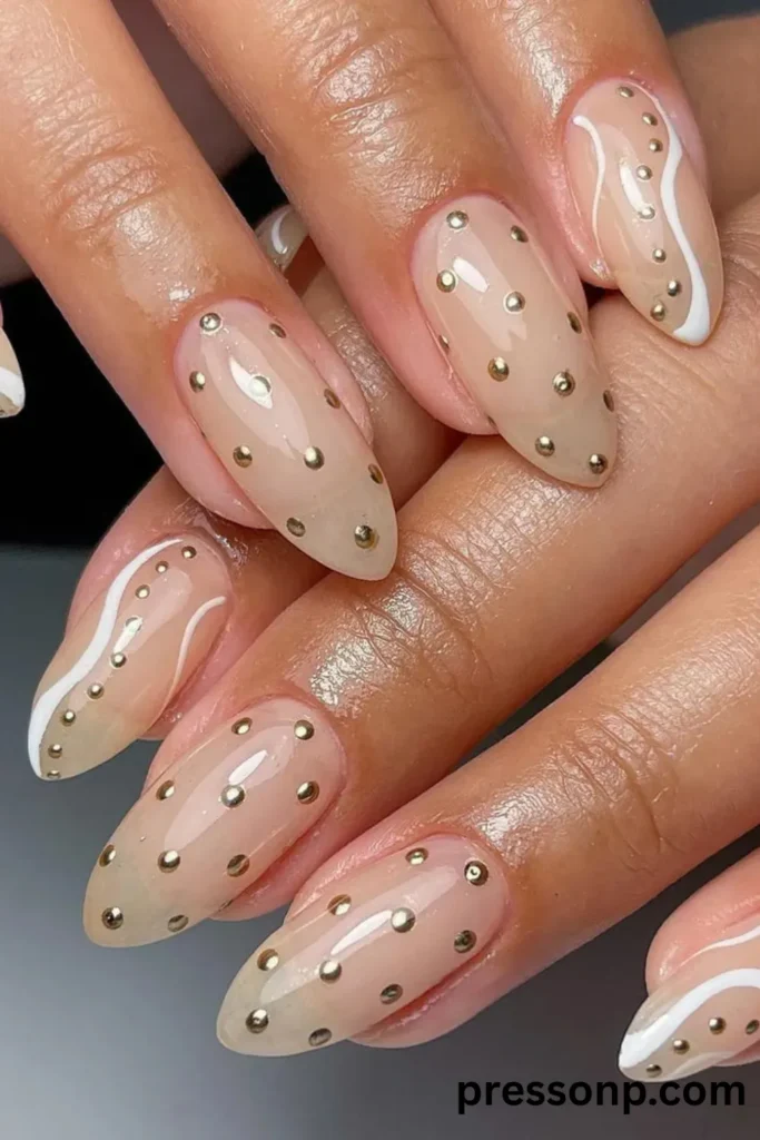 Close-up of long almond-shaped nails with a glossy, sheer nude base decorated with metallic gold studs in a polka dot pattern and abstract white wavy lines.