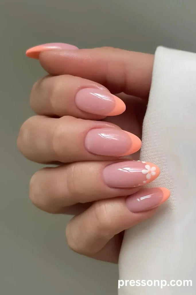 Close-up of a hand showcasing an almond-shaped manicure with a soft peach micro-French tip design and a small white floral accent on the ring finger.