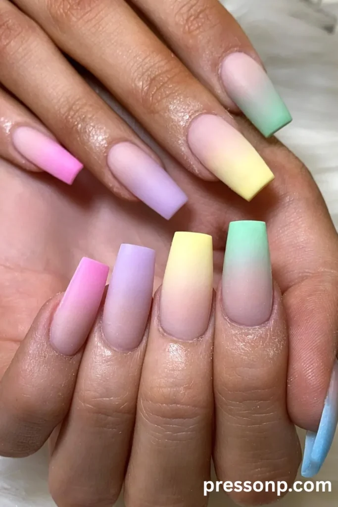 Close-up of hands with long coffin-shaped nails displaying a rainbow skittle manicure with a smooth matte ombré fade from a soft pastel base to vibrant neon tips.