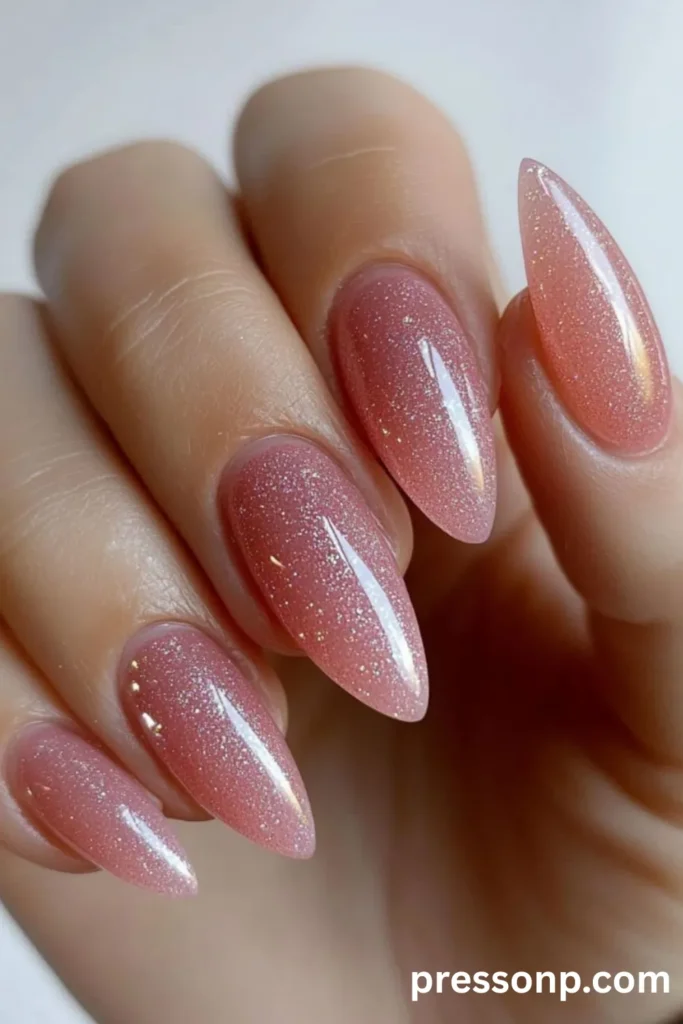 Close-up of a hand with long, almond-shaped nails featuring a glossy, shimmering shell pink polish infused with fine silver and iridescent glitter.
