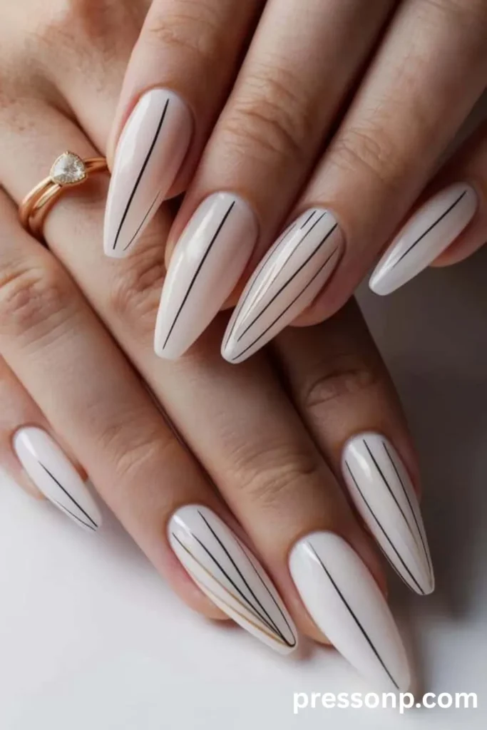 Close-up of long almond-shaped nails with a milky white base, decorated with minimalist vertical black stripes and a subtle thin gold line accent.